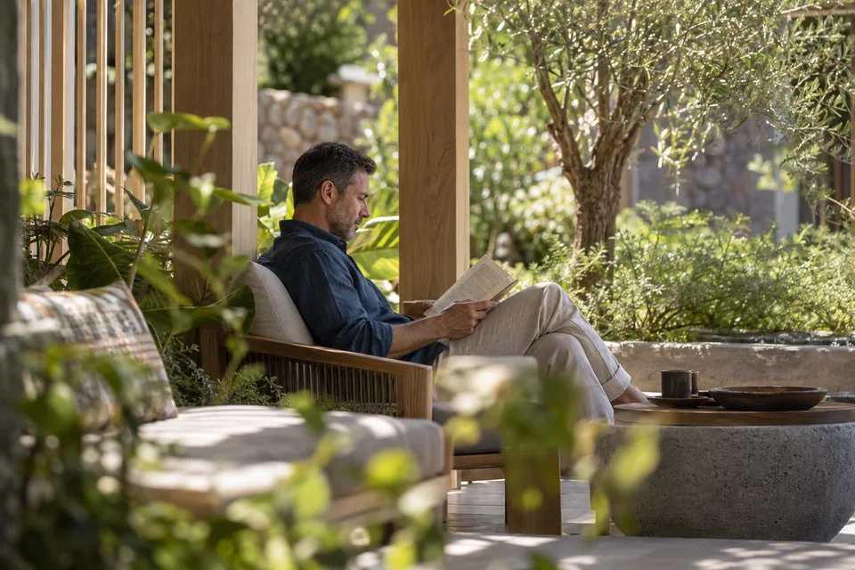 An adult reading a printed book in a shaded garden-side seating alcove.